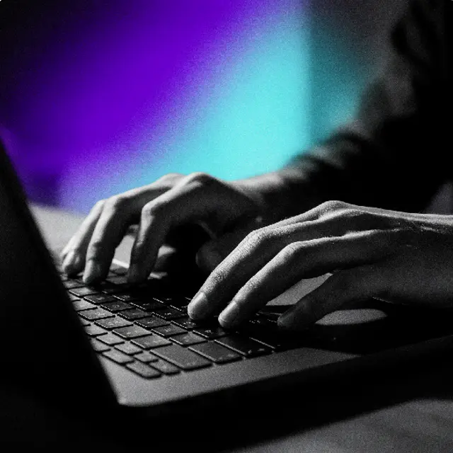 Manos escribiendo en un portátil con luz morada y cian